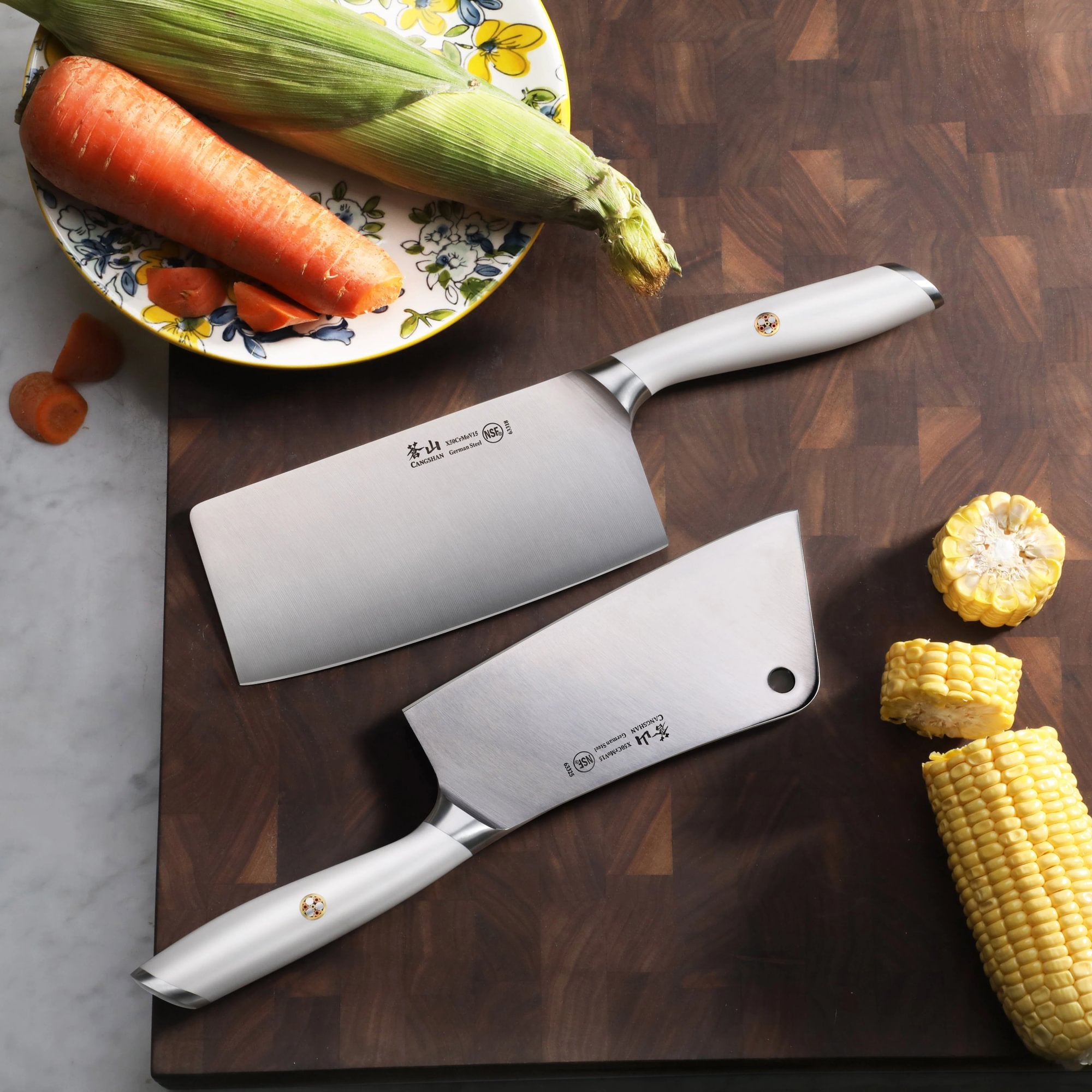 Two cleavers with white handles on a wooden cutting board next to corn, a carrot, and an ear of corn on a plate.