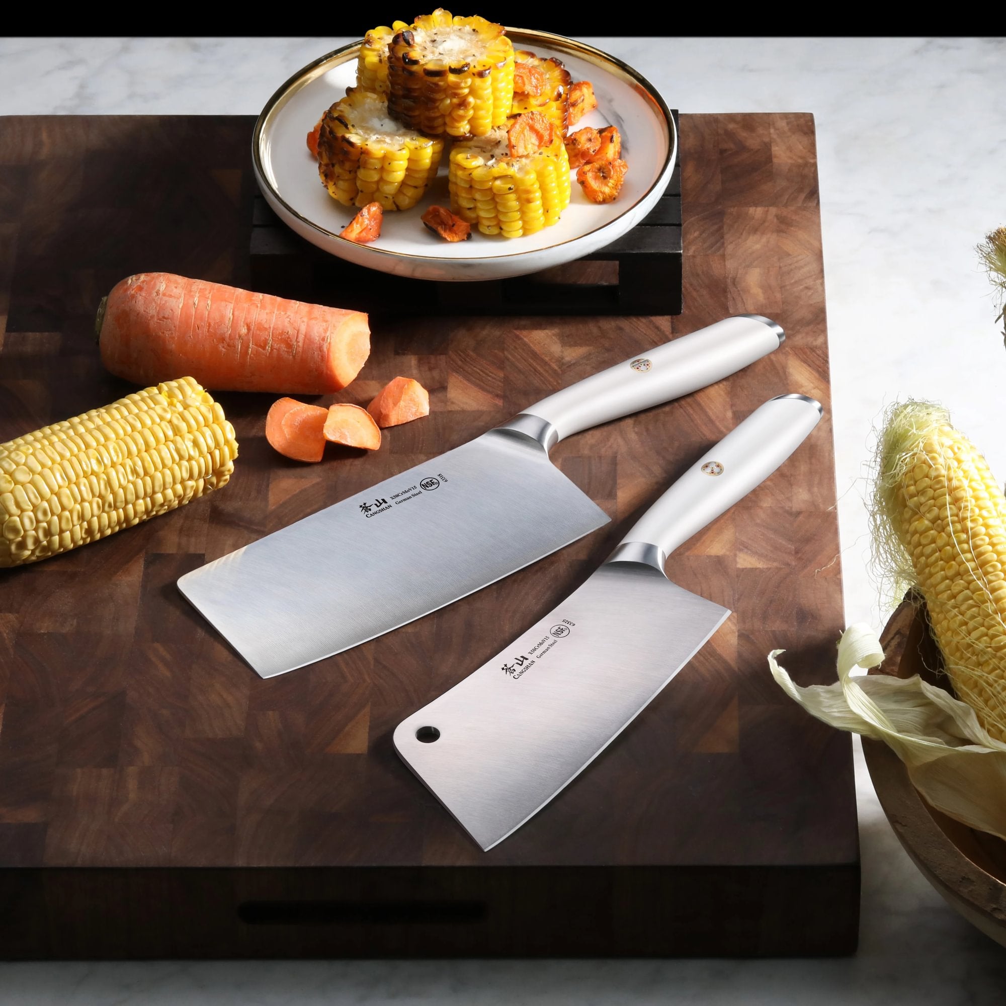 Two stainless steel cleavers on a wooden cutting board with chopped corn and carrot pieces nearby.
