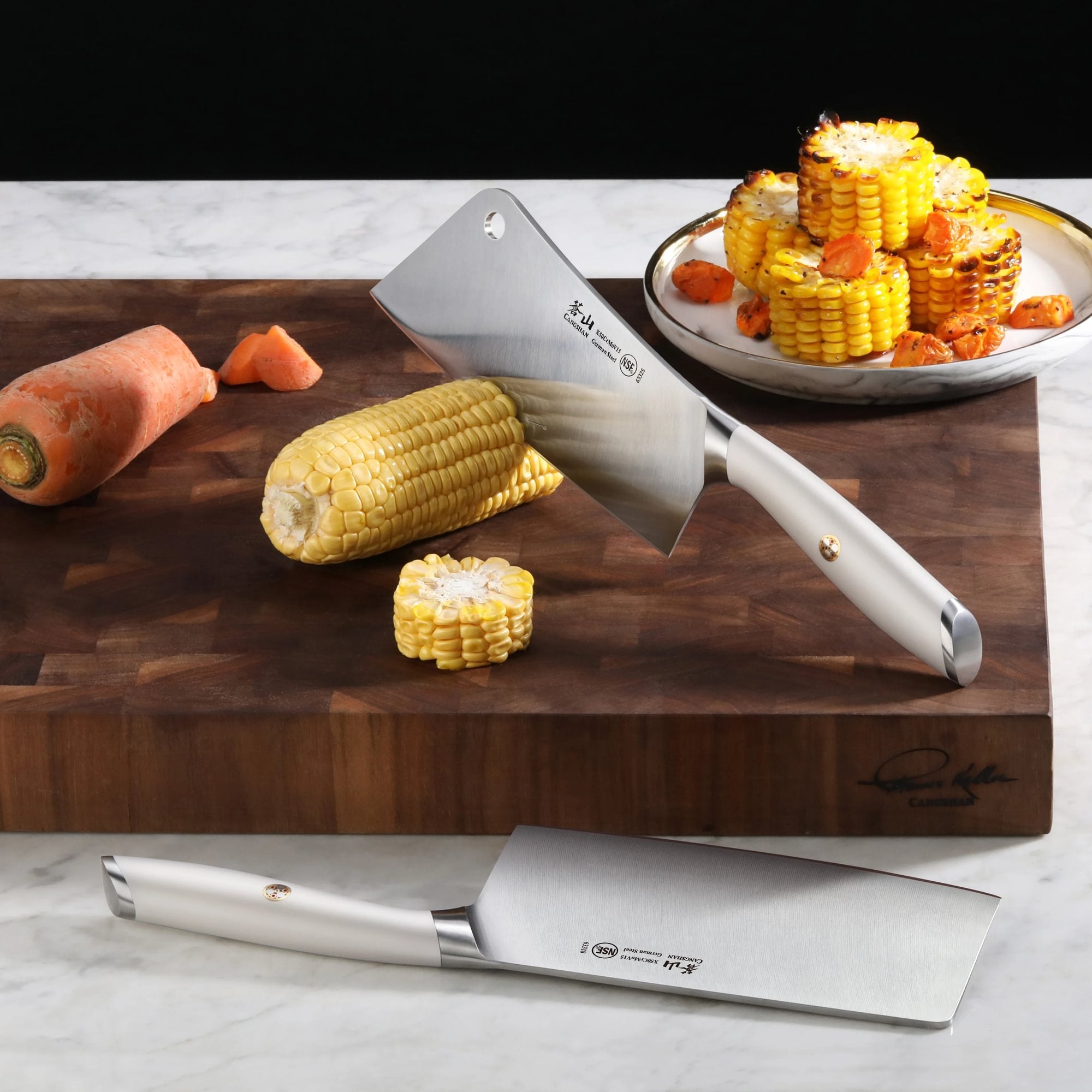 Two cleavers with silver handles on a wooden cutting board, one cutting corn, with a carrot and a plate of grilled corn and c