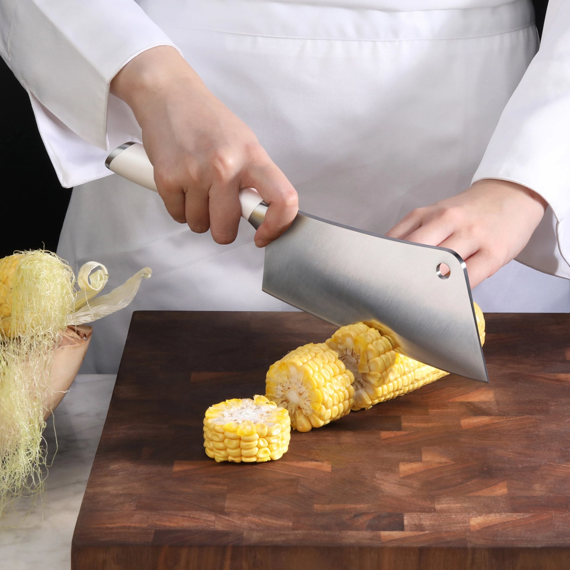 Hands holding a cleaver slicing corn on the cob on a wooden cutting board.
