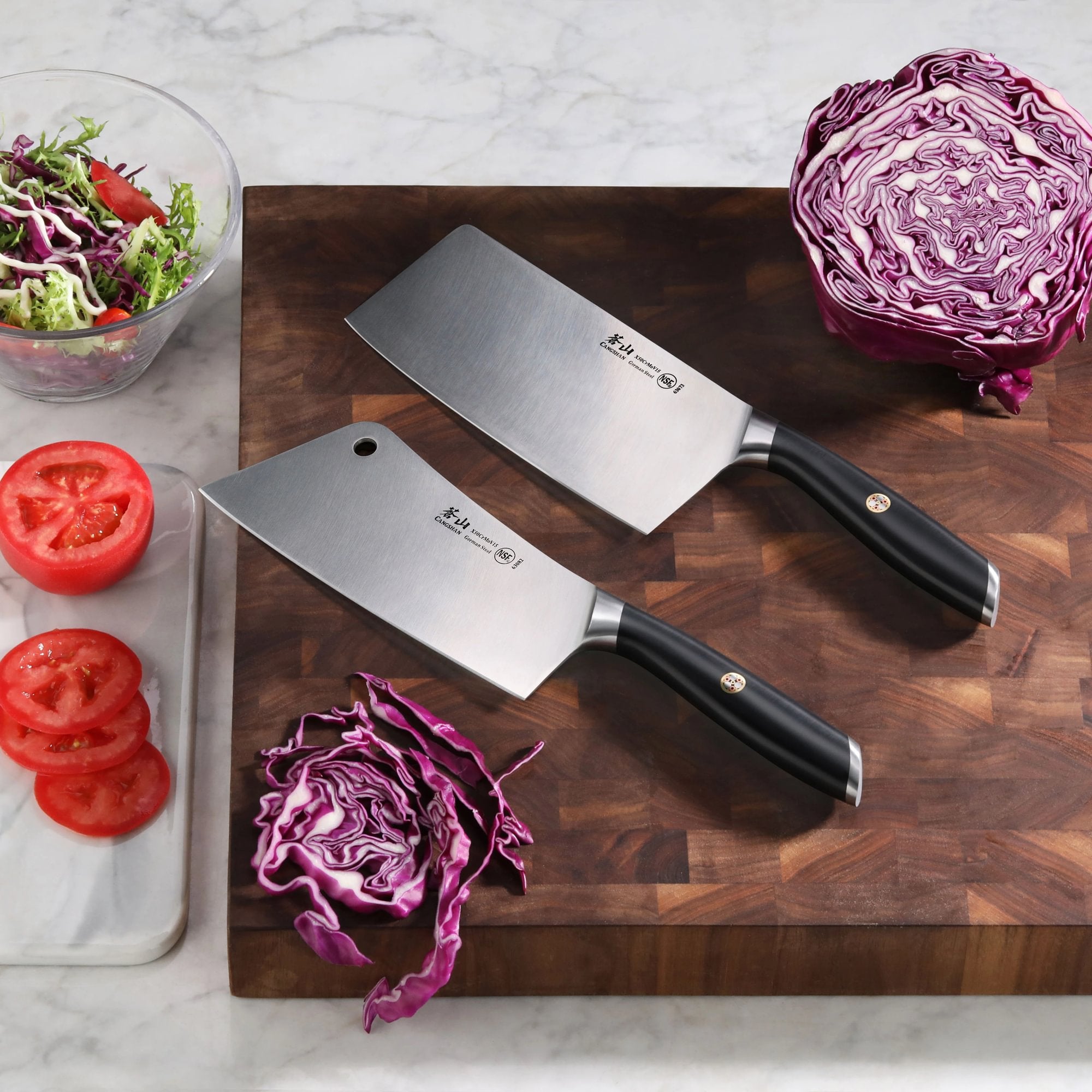 Two forged steel cleavers with black handles on a wooden cutting board, surrounded by sliced tomatoes, shredded cabbage, and