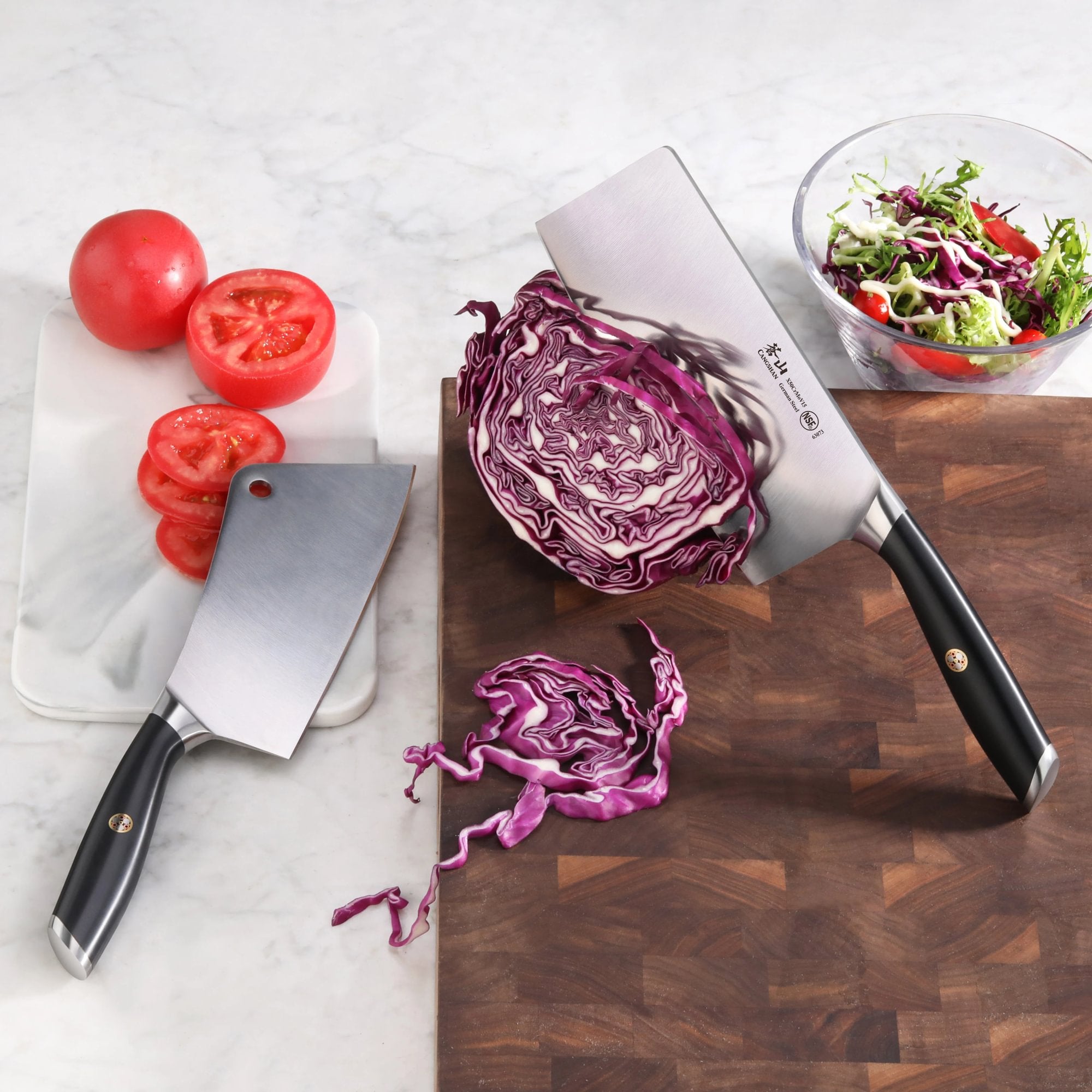 Two cleavers with black handles, one slicing red cabbage on a wooden board, the other next to sliced tomatoes on a white cutt