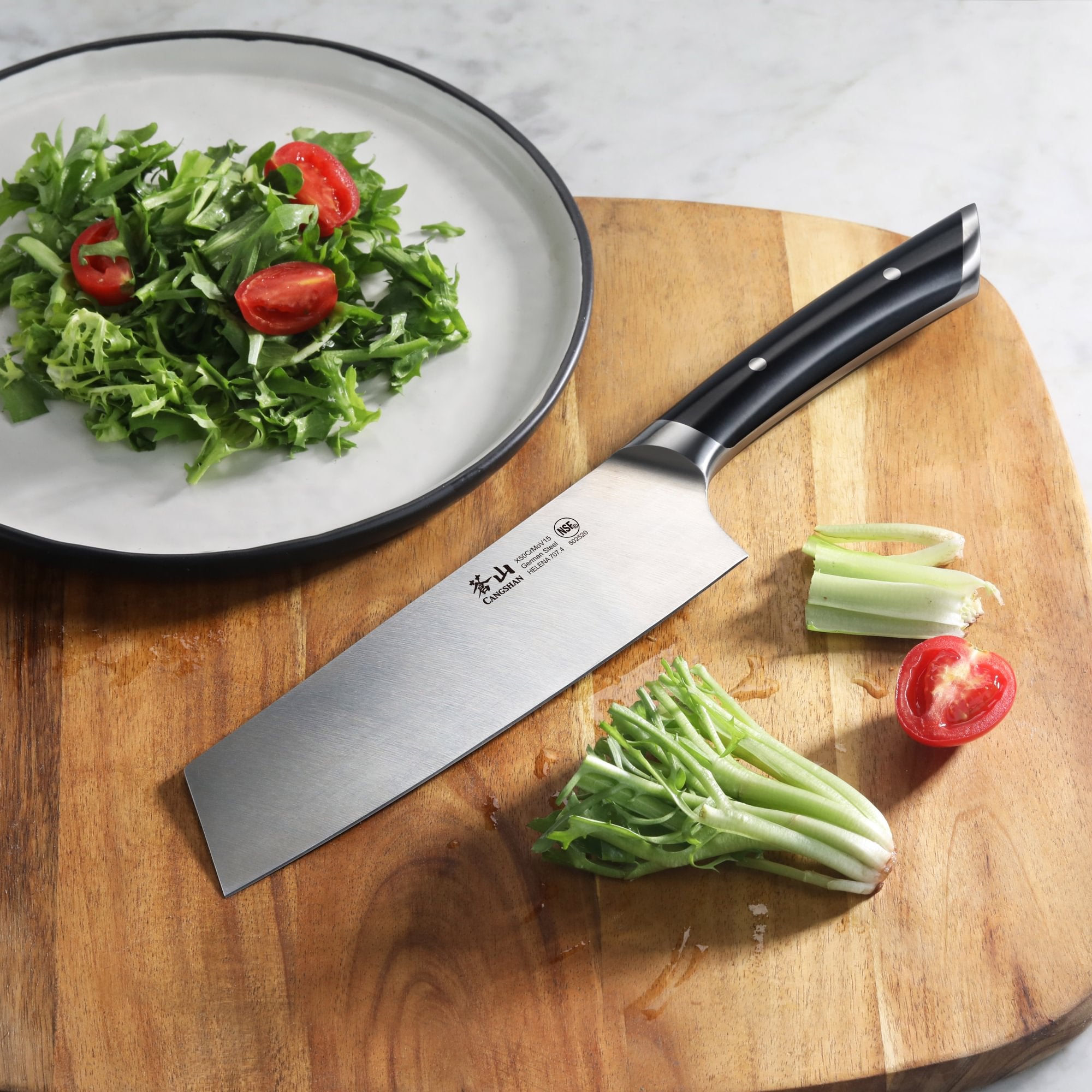 7-inch Nakiri knife with black handle on wooden cutting board next to chopped celery and tomato, plate with salad nearby.