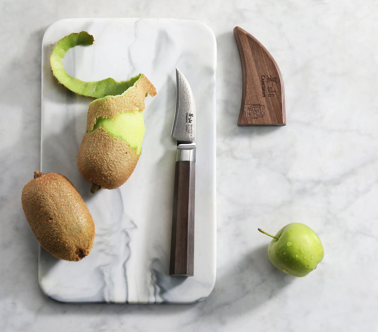 MAYA Series 2.75-inch peeling knife with sheath next to a partially peeled kiwi on a marble cutting board.