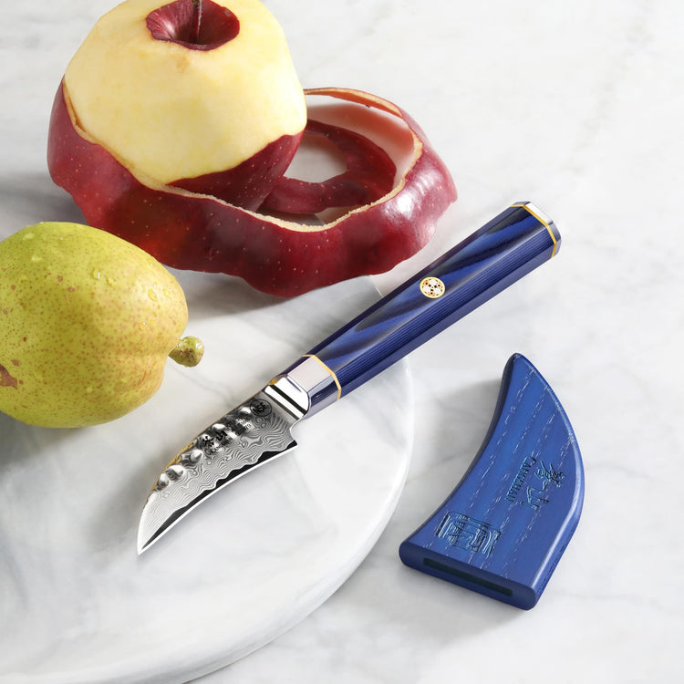 Peeling knife with blue handle and sheath next to a peeled red apple and a whole pear on a white surface.