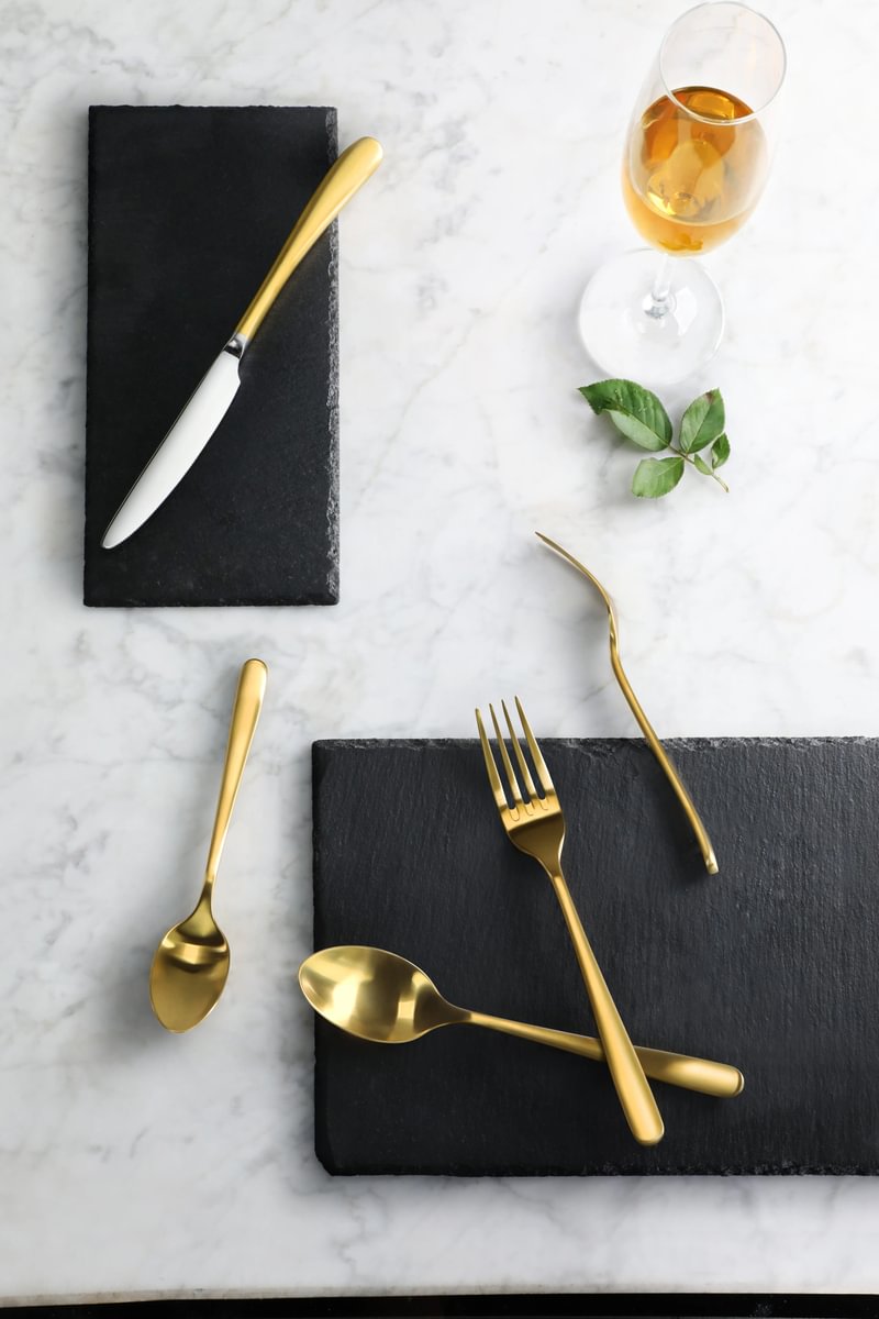 Gold RAIN II Series flatware set displayed on dark slate tiles over a marble surface.