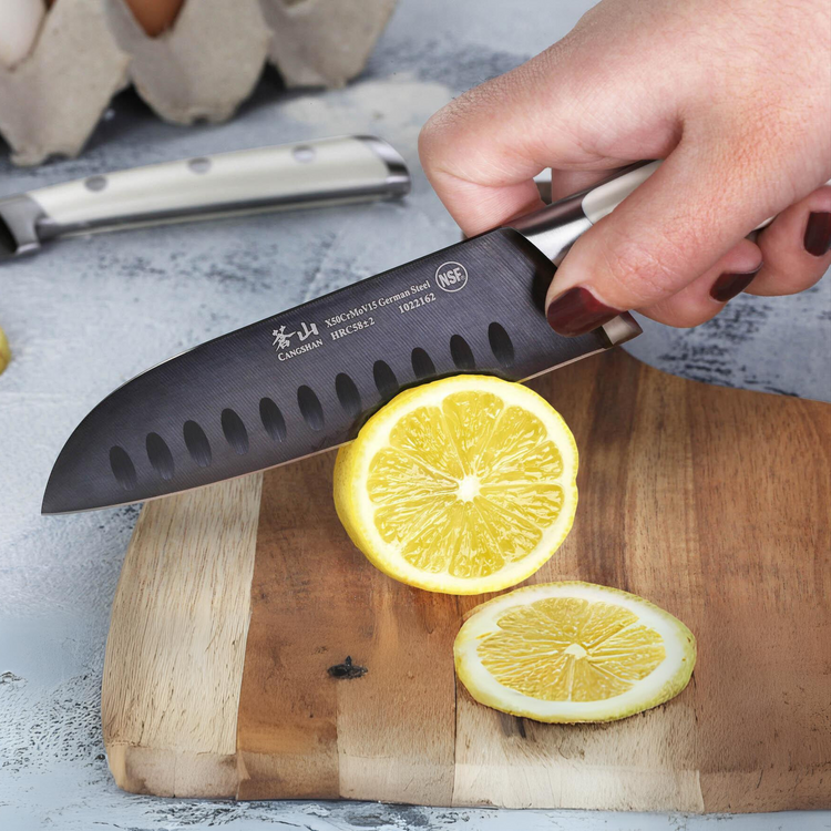 Hand holding a black knife slicing a lemon on a wooden cutting board.