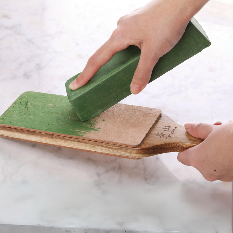 Hand applying green honing polish compound bar onto a wooden paddle with leather surface.