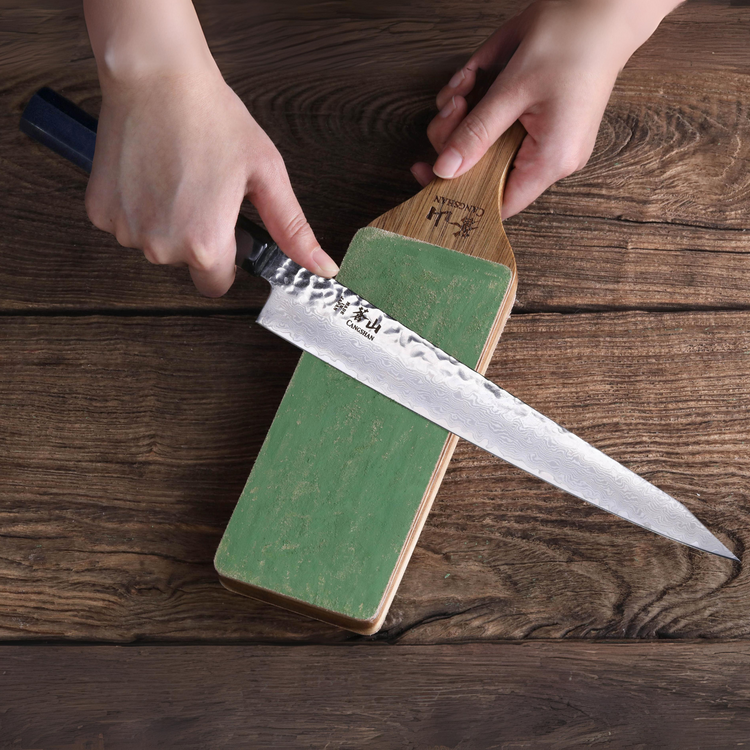 Hands holding a knife and a 2-sided leather paddle strop with a green sharpening surface on a wooden table.