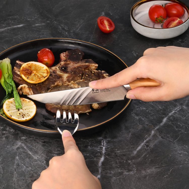 Hands using a steak knife with wooden handle to cut grilled meat on a black plate with lemon slices and vegetables.
