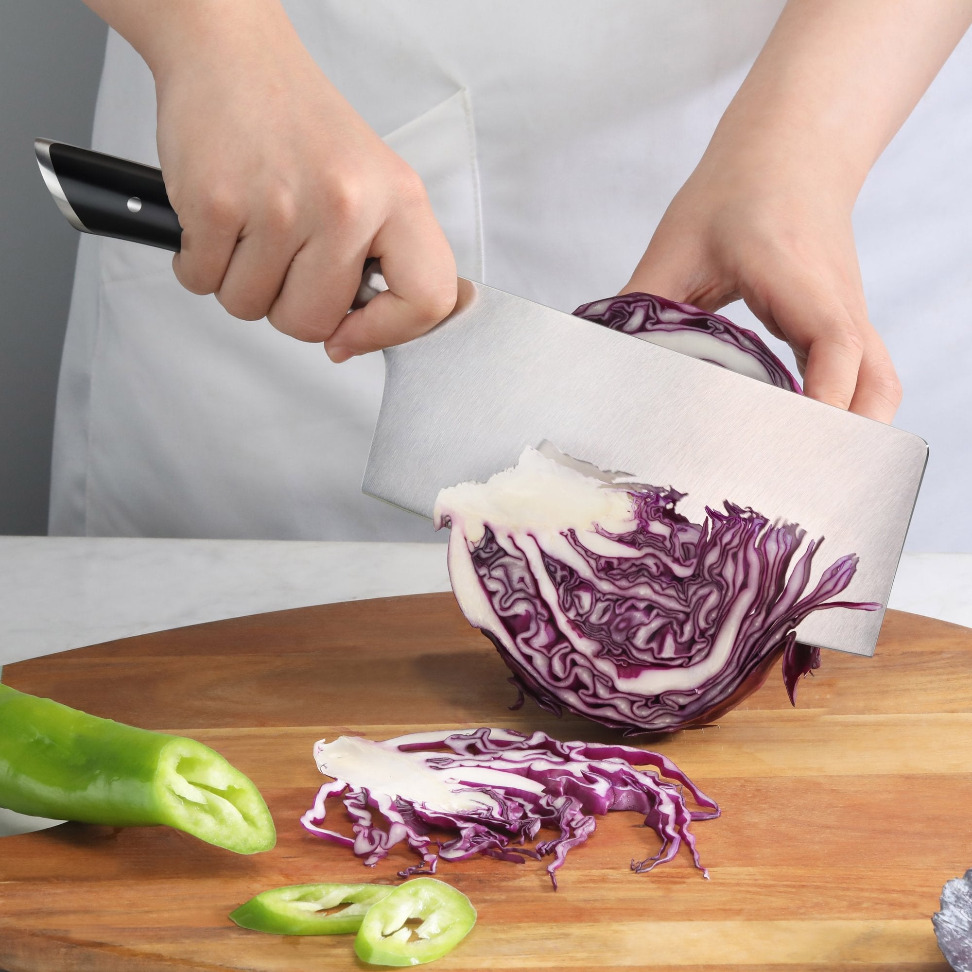 Hand holding a black-handled cleaver slicing purple cabbage on a wooden cutting board with green chili peppers nearby.