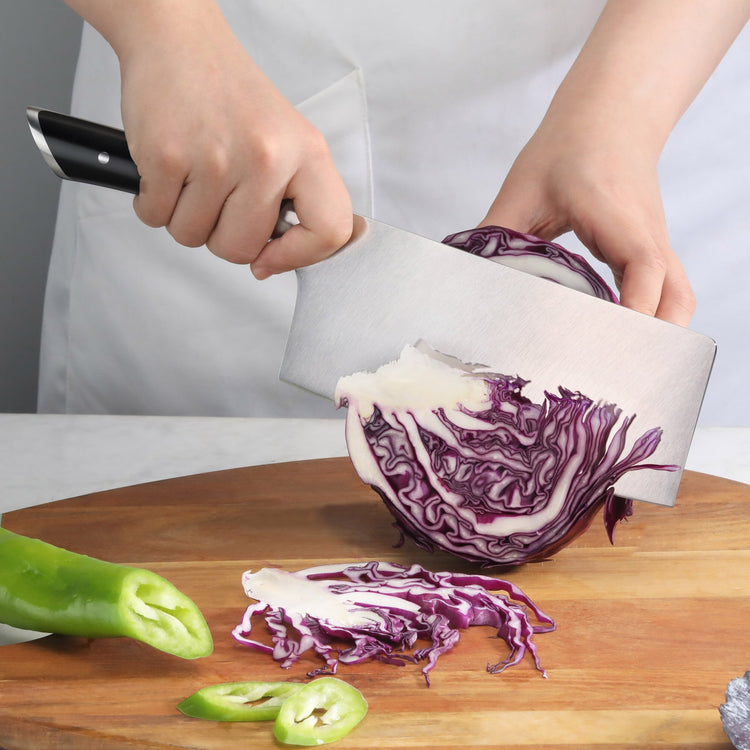 Hand holding a black-handled cleaver slicing purple cabbage on a wooden cutting board with green chili peppers nearby.
