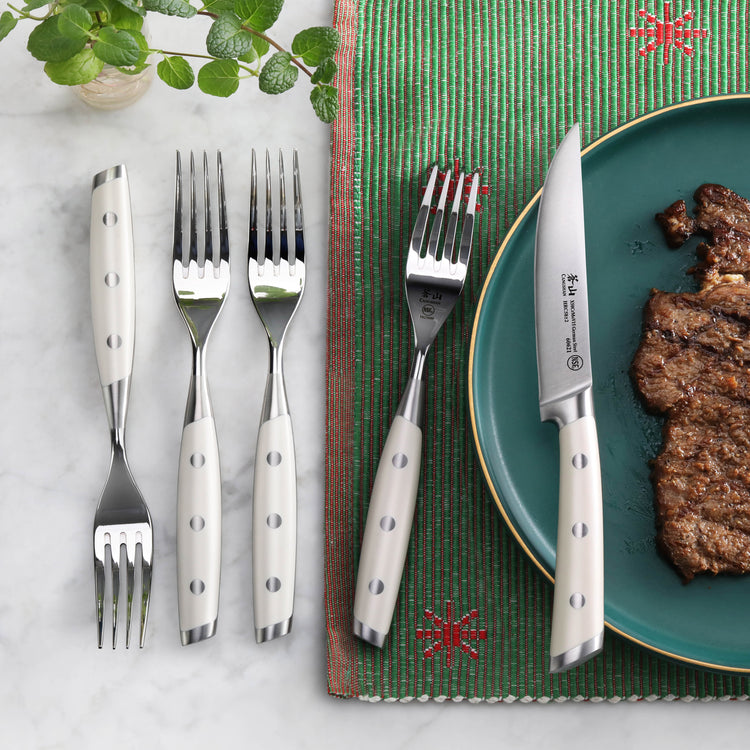 Four stainless steel steak forks with white handles and a steak knife next to a cooked steak on a green plate.