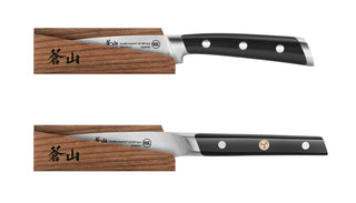 
                    
                      Two kitchen knives with black handles and three rivets each, partially inserted into solid ash wood magnetic sheaths.
                    
                  