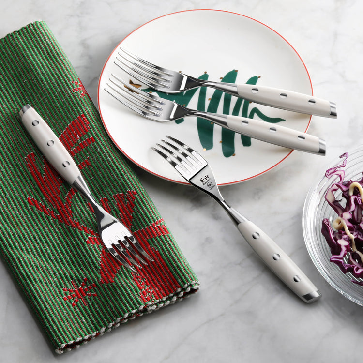 Four stainless steel steak forks with white handles arranged on a plate and green napkin on a marble surface.