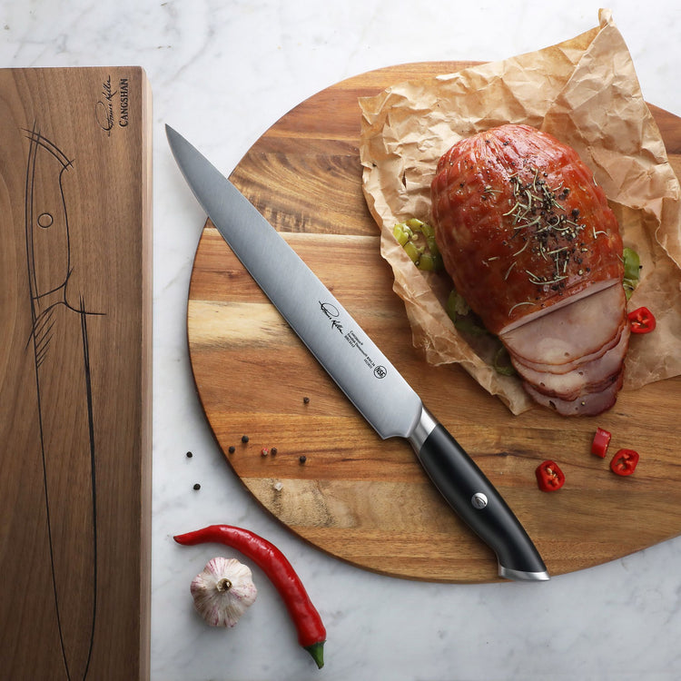 Black-handled carving knife with stainless steel blade on wooden board next to sliced roast and red chili peppers.