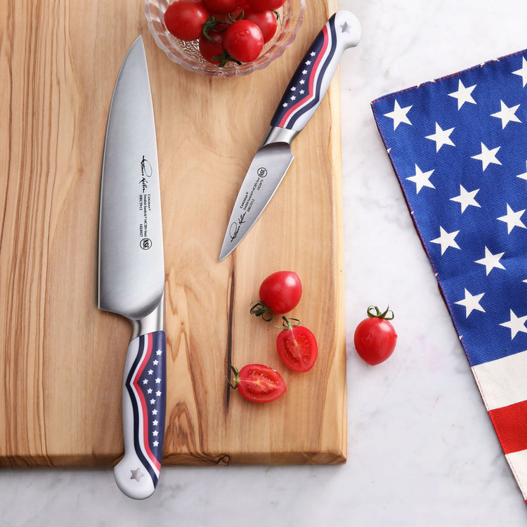 Two knives with American flag design handles on a wooden cutting board, next to halved cherry tomatoes and a star-patterned c