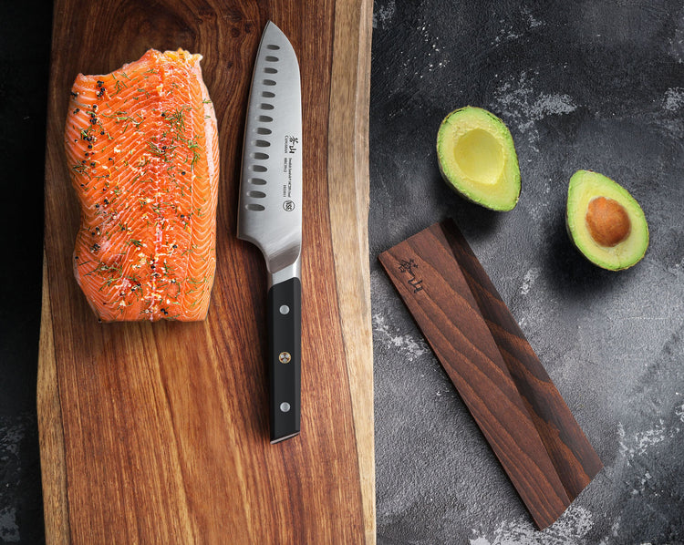Solid ash wood magnetic knife sheath next to a kitchen knife, a seasoned salmon fillet on a wooden board, and two avocado hal