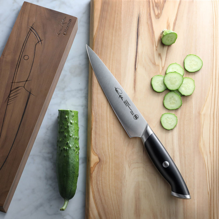 7-inch utility knife with black handle on wooden cutting board next to sliced cucumber and wooden knife box.
