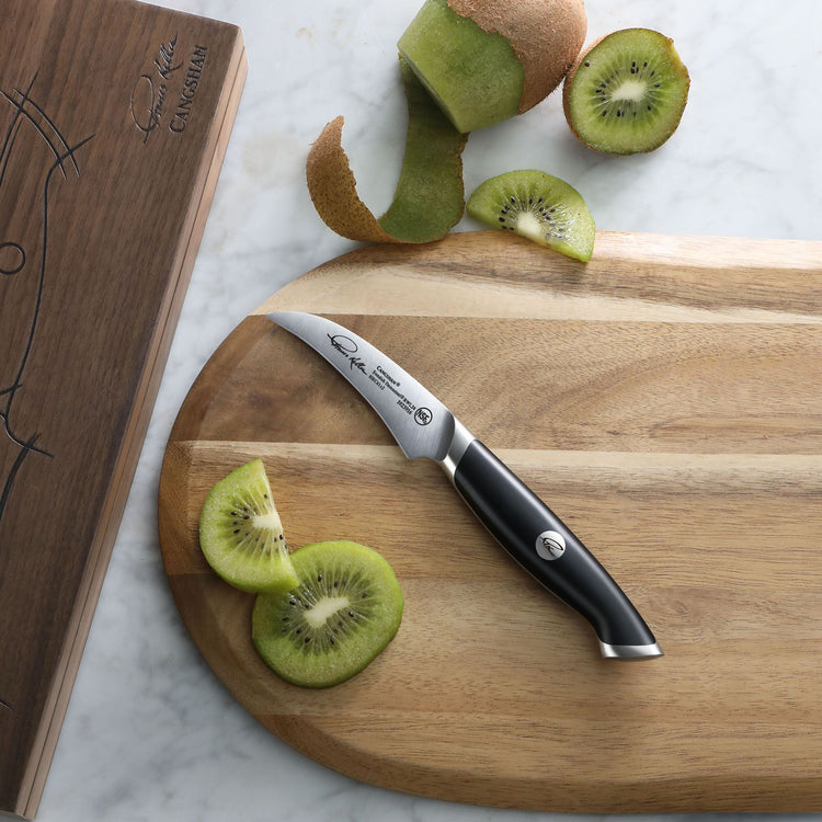 Small curved peeling knife with black handle on wooden cutting board next to peeled and sliced kiwi fruit.