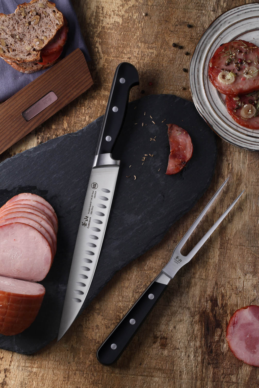 Slicing knife and meat fork on slate board with sliced ham, bread, and marinated meat on wooden table.