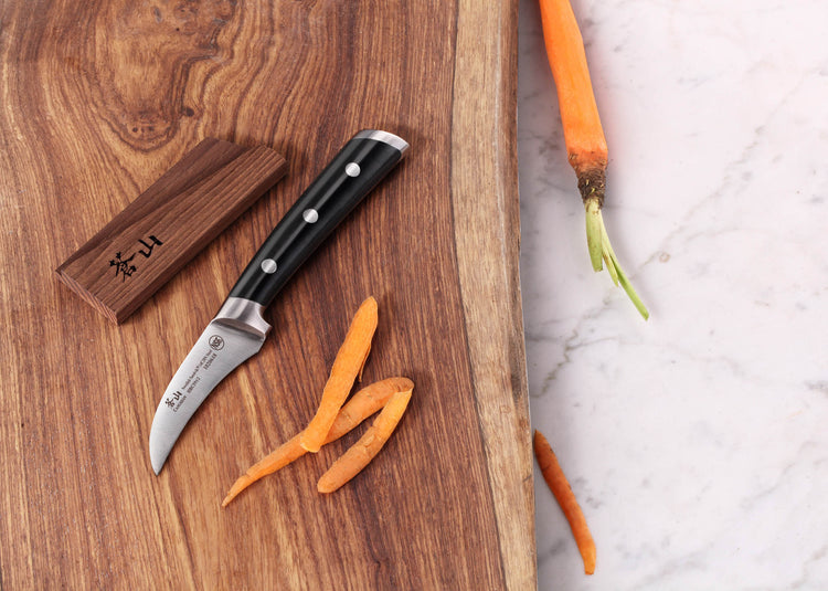 Small peeling knife with black handle and curved blade on wooden board next to peeled carrot strips and wooden sheath.