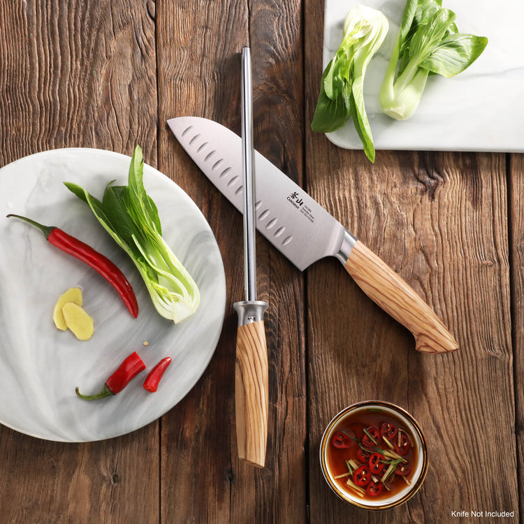 High-carbon honing steel with olivewood handle beside a chef's knife, bok choy, chili peppers, and dipping sauce on wood tabl