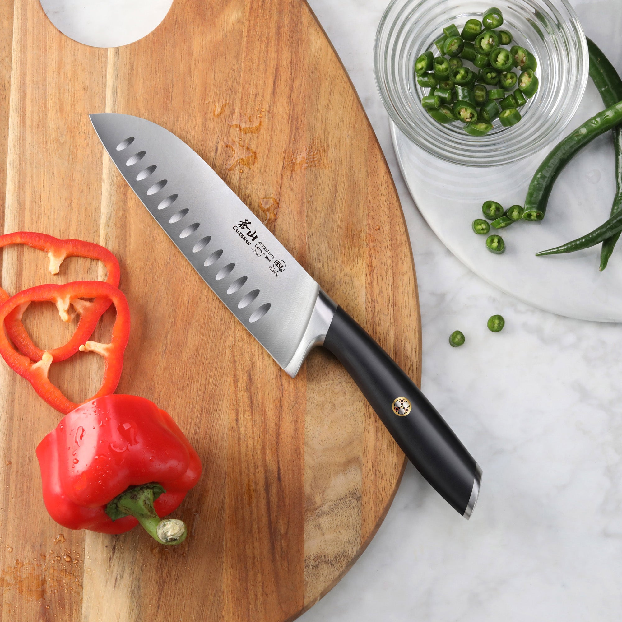 Santoku knife with black handle on wooden cutting board next to sliced red bell pepper and green chili slices.