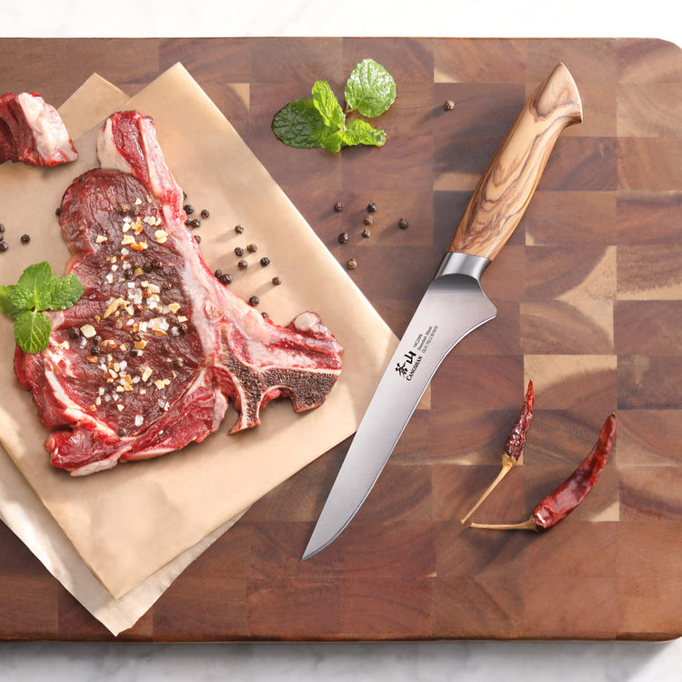 Boning knife with olivewood handle on wooden cutting board next to raw seasoned steak and dried chili peppers.