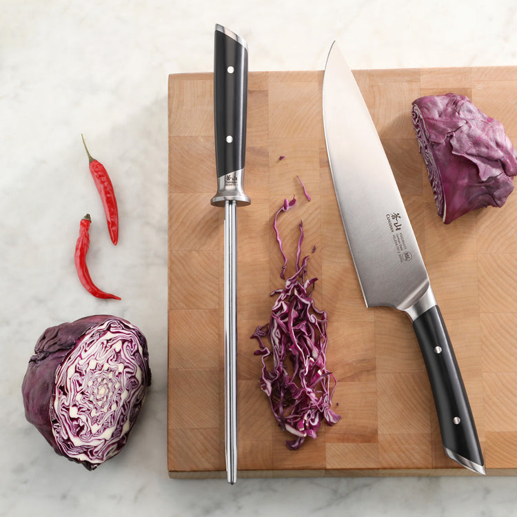 8-inch honing steel and chef's knife with black handles on a wooden cutting board, surrounded by red cabbage and chili pepper
