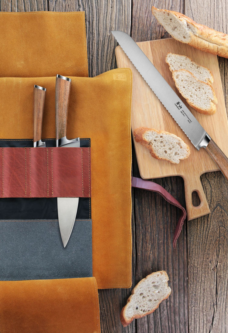 Three knives with wooden handles in a leather roll knife case next to a serrated knife cutting sliced bread on a wooden board