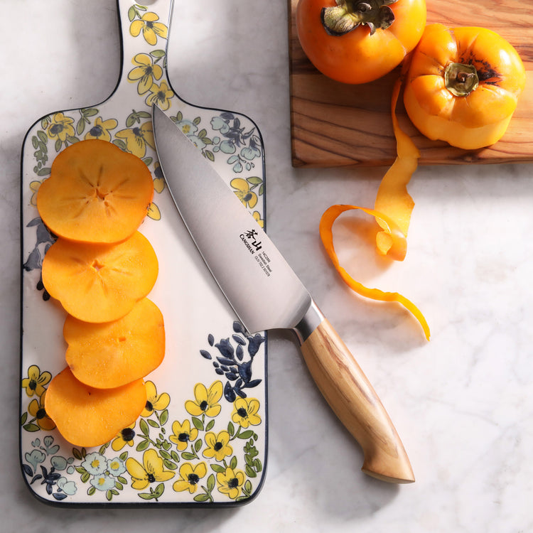 6-inch chef's knife with wooden handle slicing a peeled persimmon on a floral cutting board next to whole persimmons.