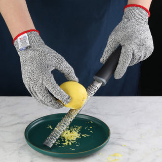 
                    
                      Hands wearing gray cut resistant gloves grating a lemon over a green plate on a marble surface.
                    
                  