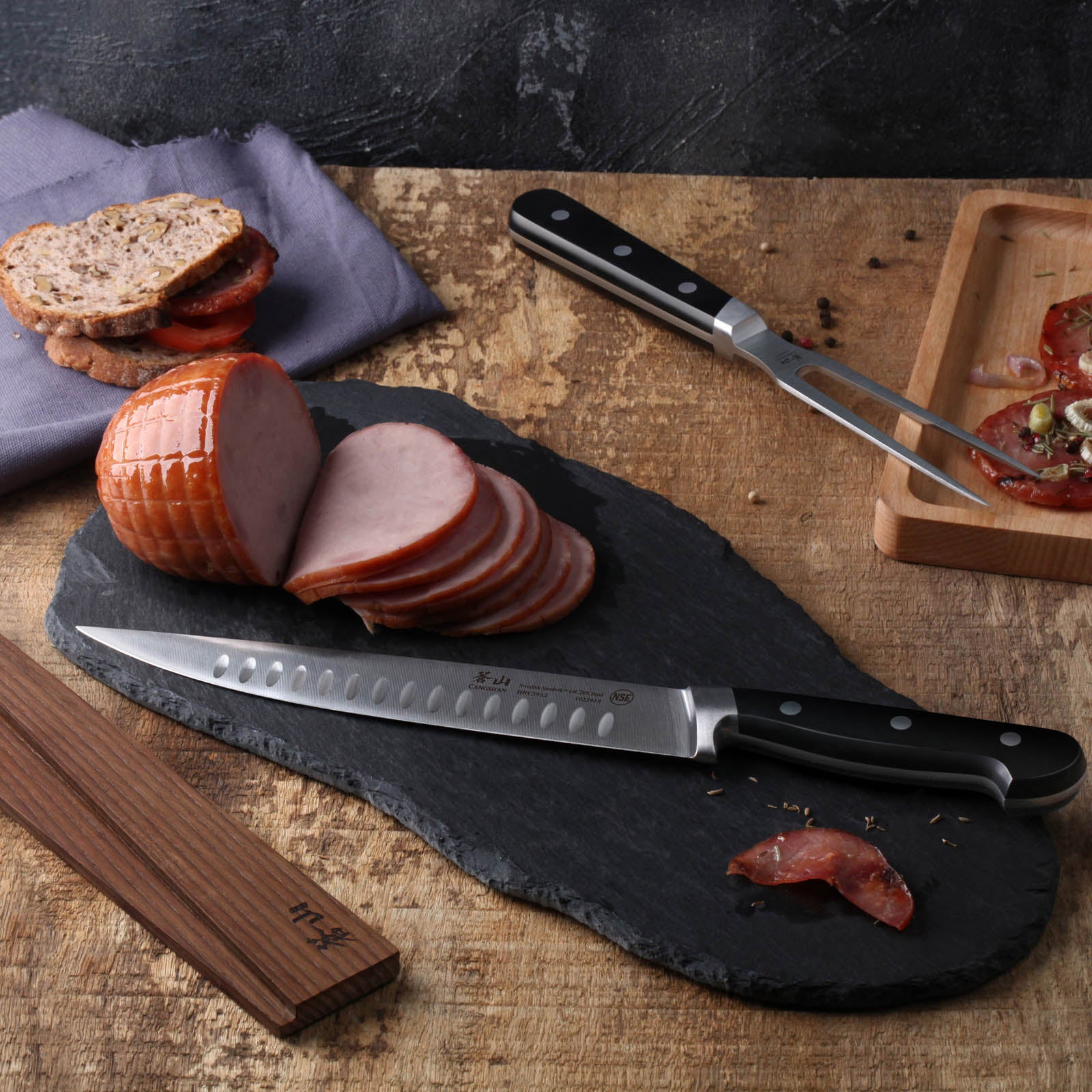 Sliced ham on a black slate board with a serrated knife, sandwich, meat fork, and wooden tray with sliced meat.
