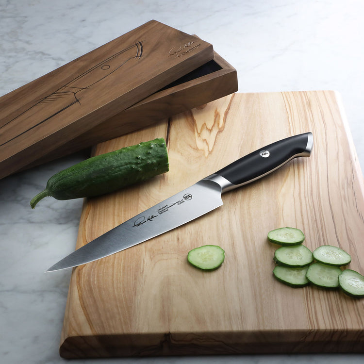 7-inch utility knife with black handle on wooden cutting board, partially sliced cucumber beside it, wooden knife box nearby.