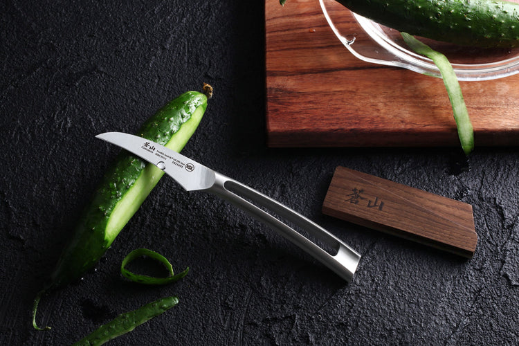 TN1 Series 2.75-inch peeling knife with curved blade and metal handle slicing a cucumber on a black surface next to an ash wo