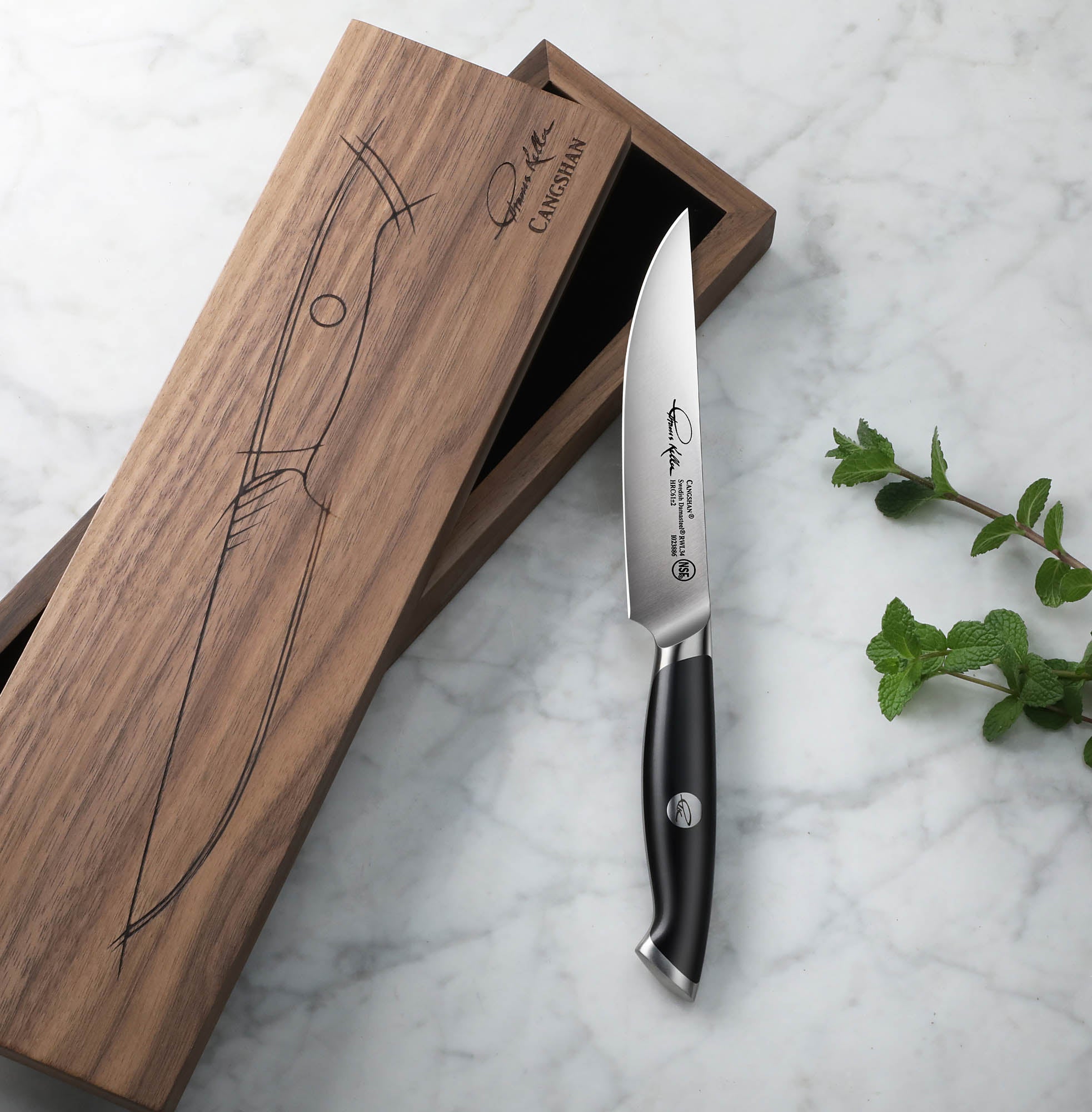 Black-handled steak knife with polished blade next to a wooden box with knife illustration on marble surface.