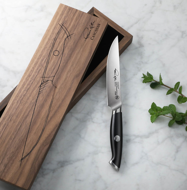 Black-handled steak knife with polished blade next to a wooden box with knife illustration on marble surface.