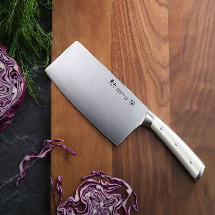 Cleaver knife with white handle on wooden cutting board next to sliced purple cabbage and green herbs.