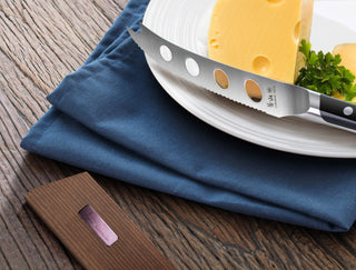 
                    
                      Solid ash wood magnetic knife sheath placed on a wooden surface next to a plate with cheese and a serrated knife.
                    
                  