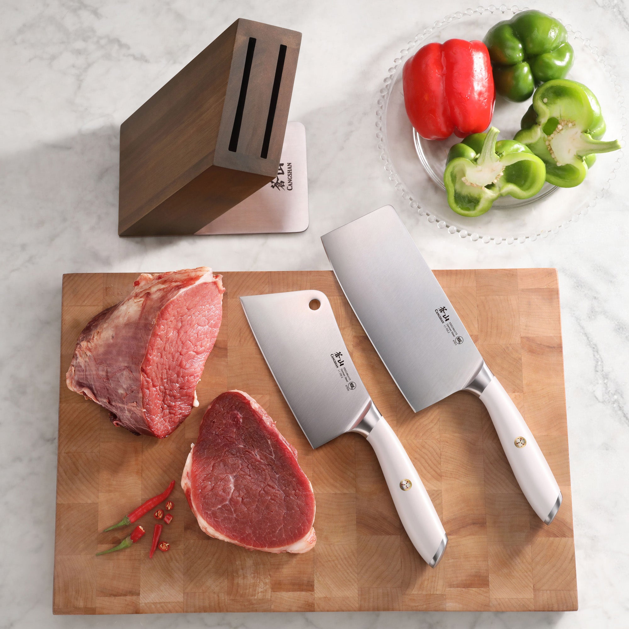 Three cleaver knives with white handles on a wooden cutting board with raw meat and chili peppers, next to an empty wooden kn