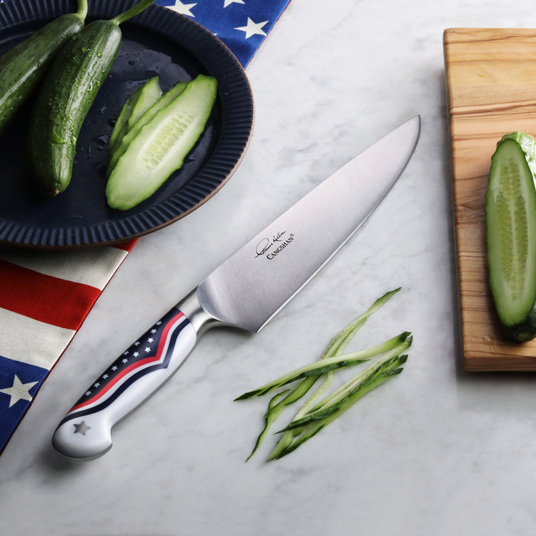 Chef's knife with American flag design handle next to sliced cucumber on marble surface and wooden cutting board.