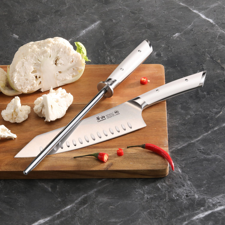 8-inch honing steel and chef's knife with white handles on wooden cutting board with cauliflower and red chili peppers.