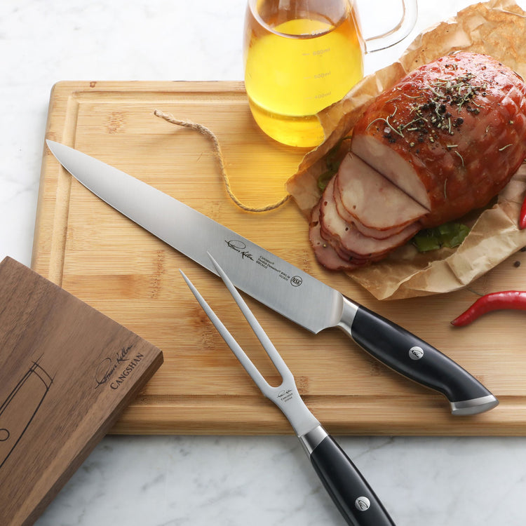Carving knife and fork with black handles on wooden board next to sliced roast and glass pitcher of oil.