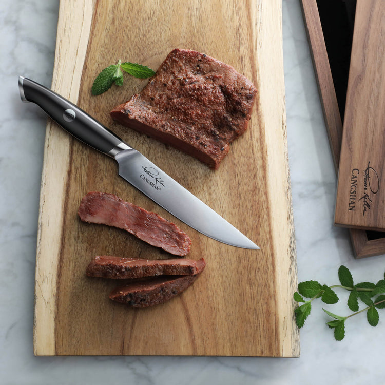 TKSC 5-inch steak knife with black handle slicing cooked steak on wooden board with mint garnish.