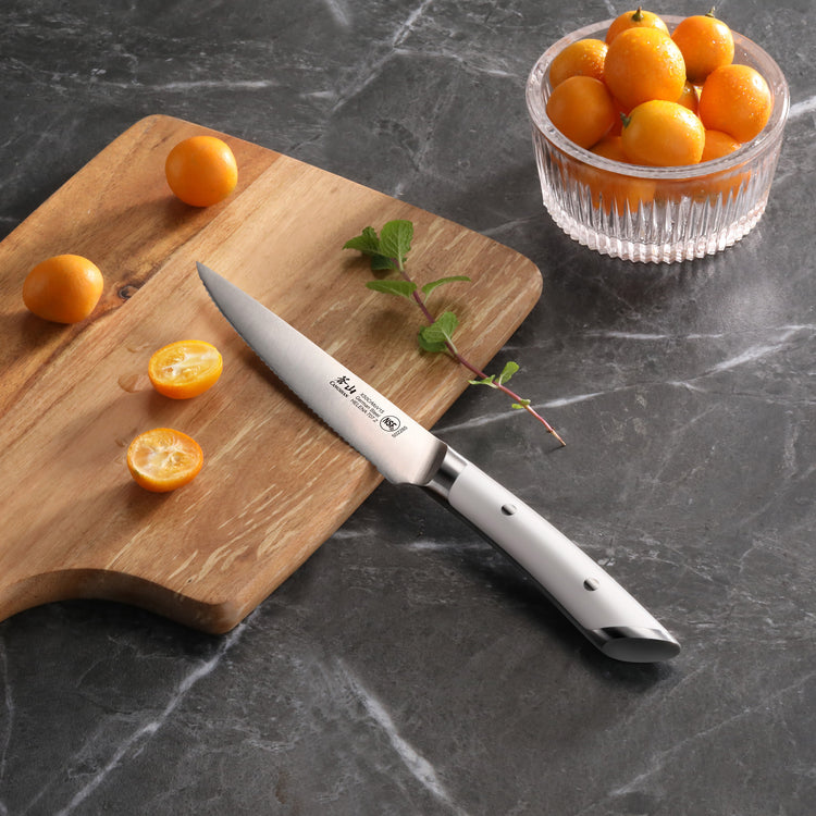 Serrated utility knife with stainless steel blade on wooden cutting board next to halved kumquats and a glass bowl of kumquat