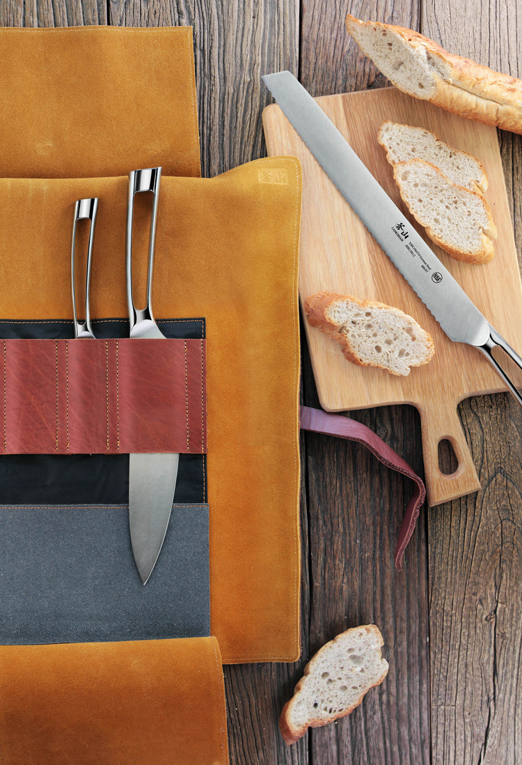Four knives with silver handles in a leather roll knife set next to a cutting board with sliced bread.