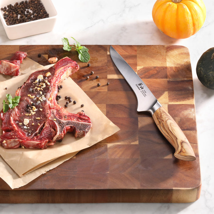 Boning knife with olivewood handle on wooden cutting board next to raw seasoned steak and peppercorns.