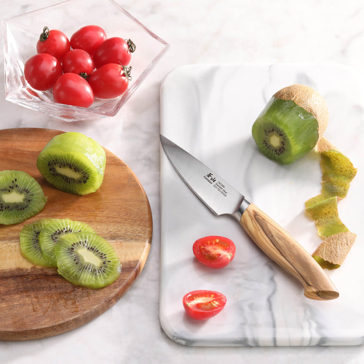 Paring knife with olivewood handle peeling a kiwi, sliced kiwi on wooden board, and cherry tomatoes in glass bowl.
