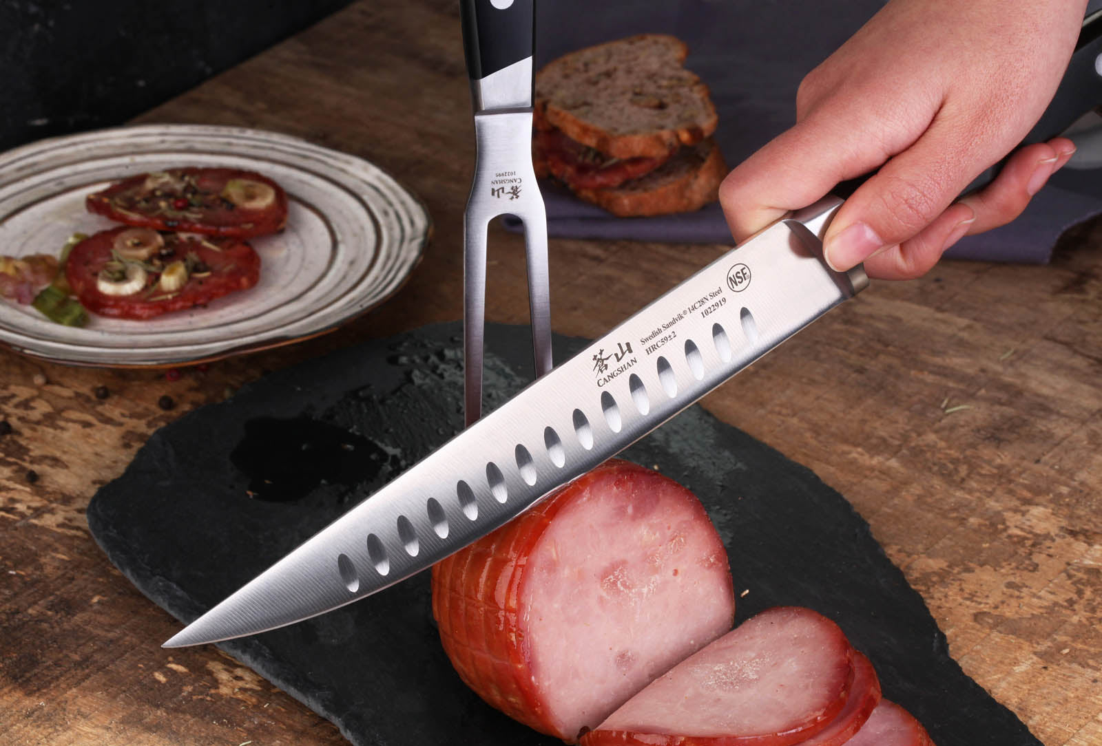 Hand holding a knife slicing cooked ham on a black slate board with a fork and plate of sliced tomatoes in the background.
